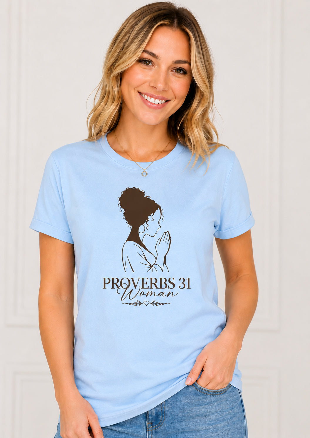 A smiling blonde woman wearing a light blue graphic t-shirt with a black silhouette of a praying woman and the text 'PROVERBS 31 Woman' printed on the front, paired with blue jeans.