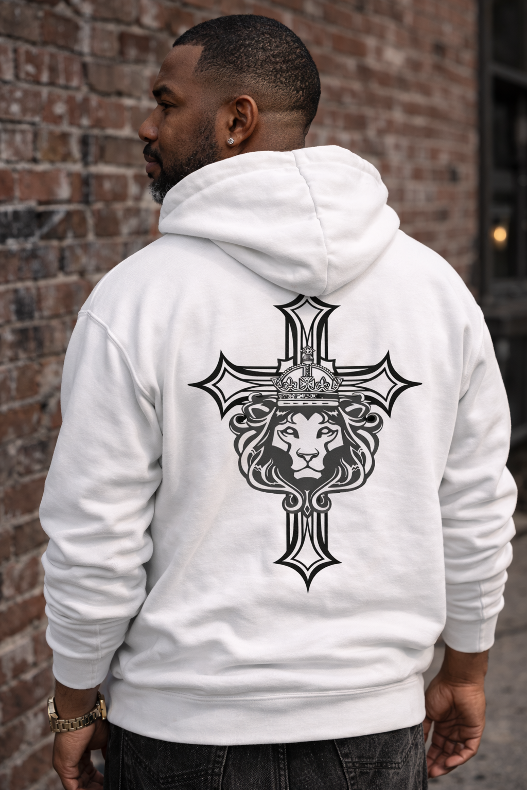 Man wearing a white hoodie with a black lion and cross design on the back, standing against a brick wall.